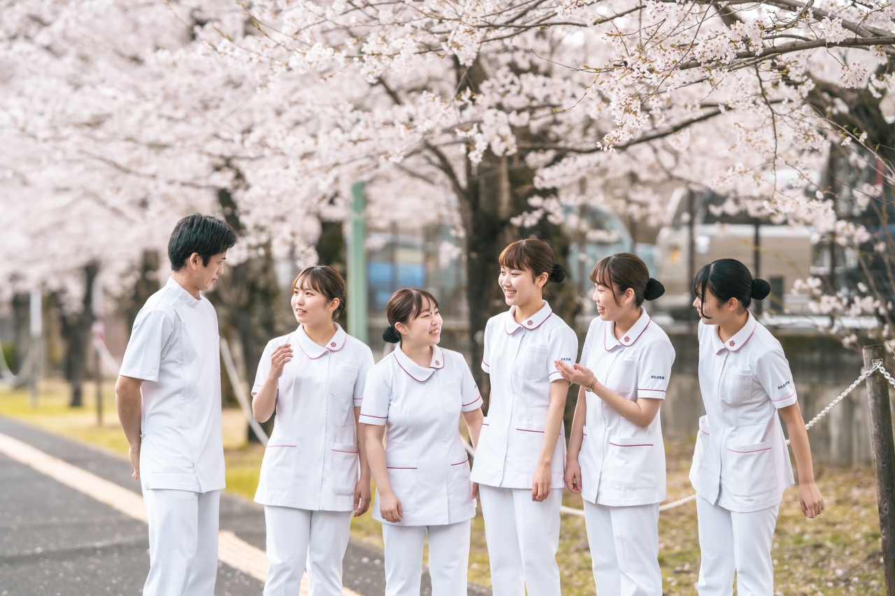 4月学校と病院をつなぐ道に桜が満開。その下を学生が楽しそうにおしゃべりしている